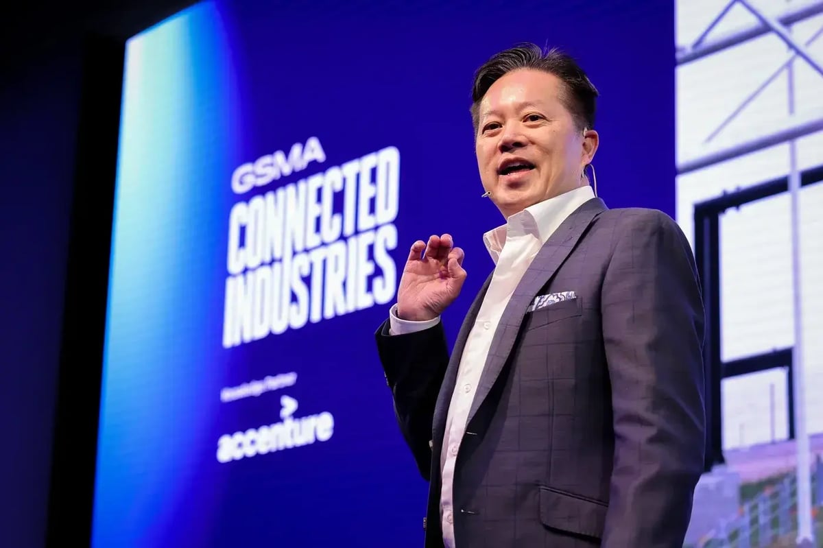 A speaker in a dark suit and white shirt presents on stage at MWC25's Connected Industries session. He is gesturing while speaking, with a microphone headset. Behind him, a large screen displays the event branding with the words 'GSMA Connected Industries' and 'Accenture' as a partner. The stage is illuminated with blue and white lighting.