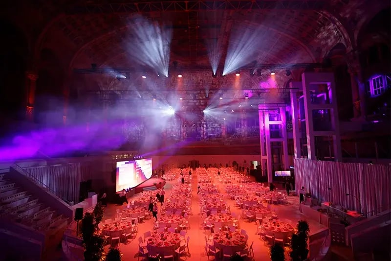 Wide-angle view of an elegant banquet hall set for a formal event, featuring rows of round dining tables with red floral centerpieces. Dramatic purple and orange stage lighting fills the grand domed ceiling, highlighting ornate architectural details and creating a theatrical atmosphere. A large screen is positioned near the stage at the front of the venue.