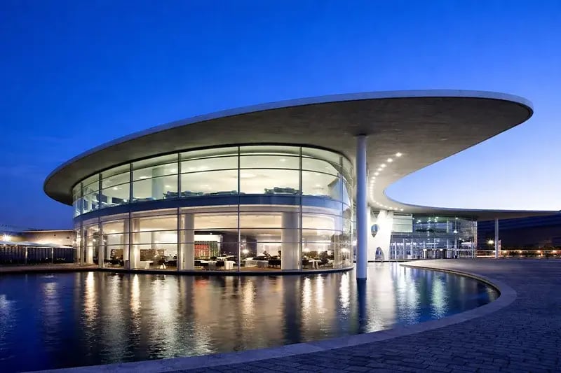 Futuristic glass building with a wide, curved roof is illuminated against a twilight sky. It overlooks a reflecting pool, evoking a sleek, modern tone.
