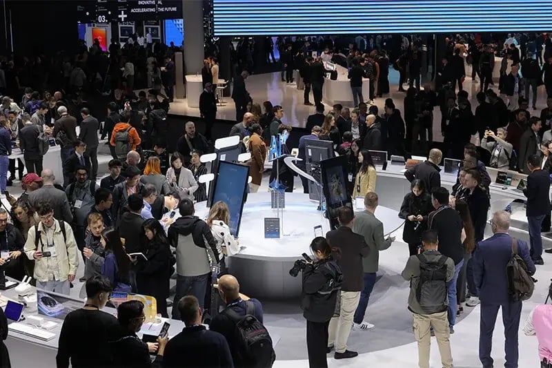 A bustling tech expo features a crowd exploring digital displays and gadgets at a large, circular booth. The atmosphere is lively and engaging.