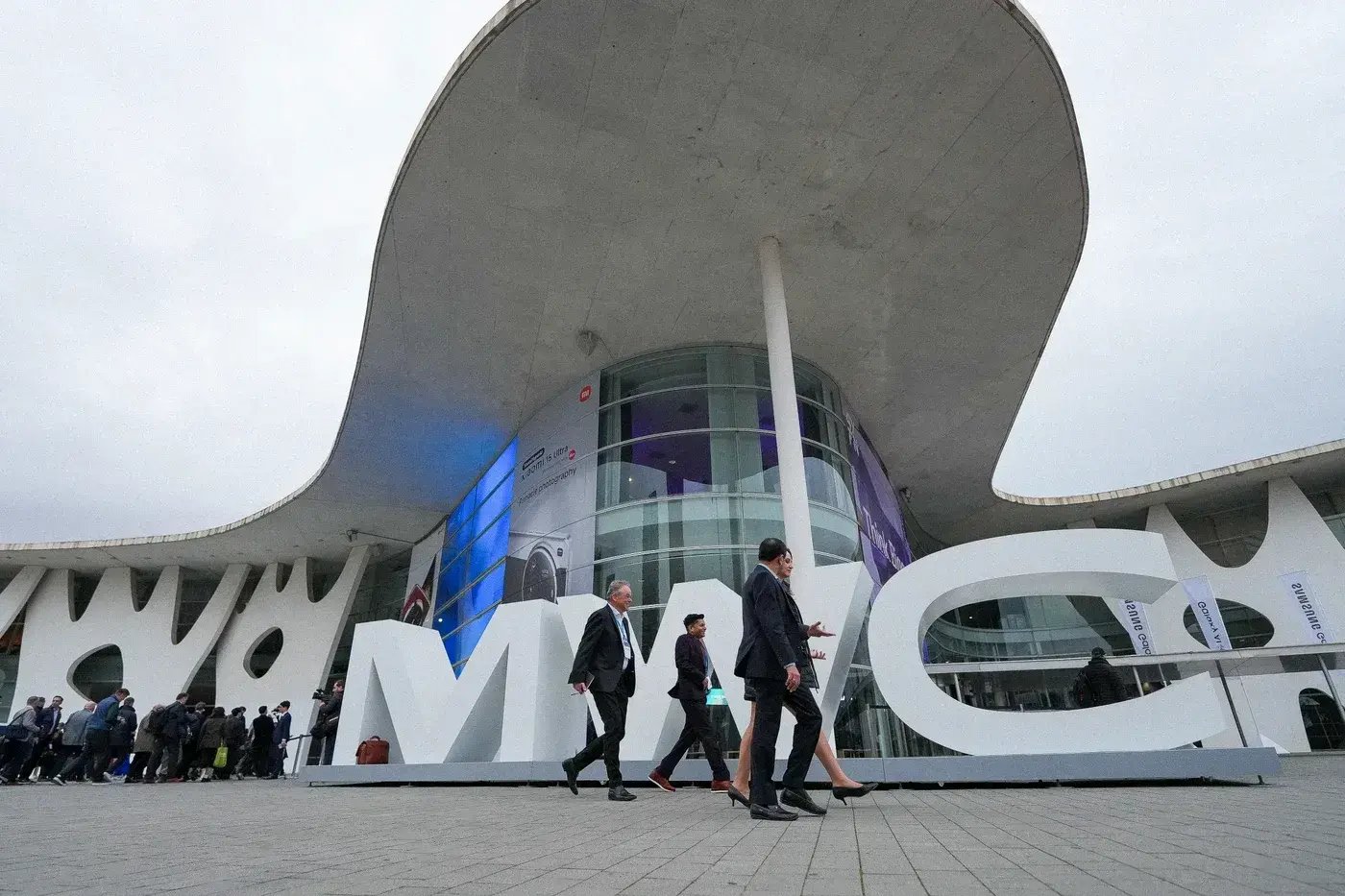 Exterior view of the MWC25 venue at Fira Barcelona, featuring the large 'MWC' sign in the foreground. Several business professionals in formal attire walk past the sign, while other attendees line up to enter the building. The venue's modern architectural design with curved structures and glass panels is visible, along with banners and advertisements promoting the event.
