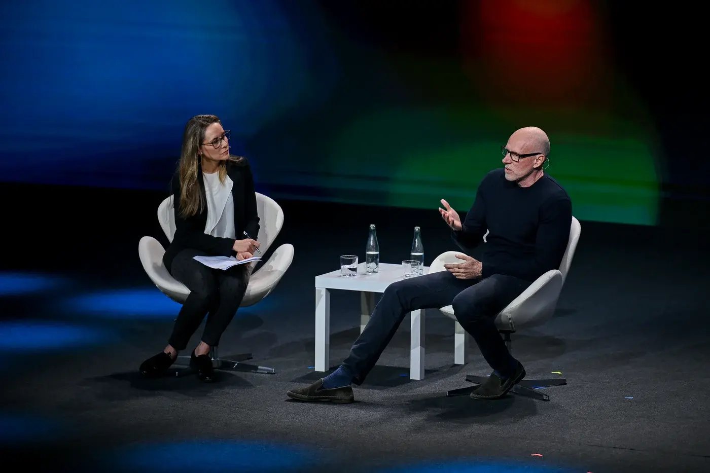 A keynote discussion at MWC25 featuring journalist Nina dos Santos and business professor Scott Galloway on a dimly lit stage with a colorful abstract background. Nina dos Santos, seated on the left, wears a black blazer and white blouse, holding a notepad while attentively listening. Scott Galloway, seated on the right, wears a black sweater and glasses, gesturing as he speaks. A small table between them holds two bottles of water and glasses.