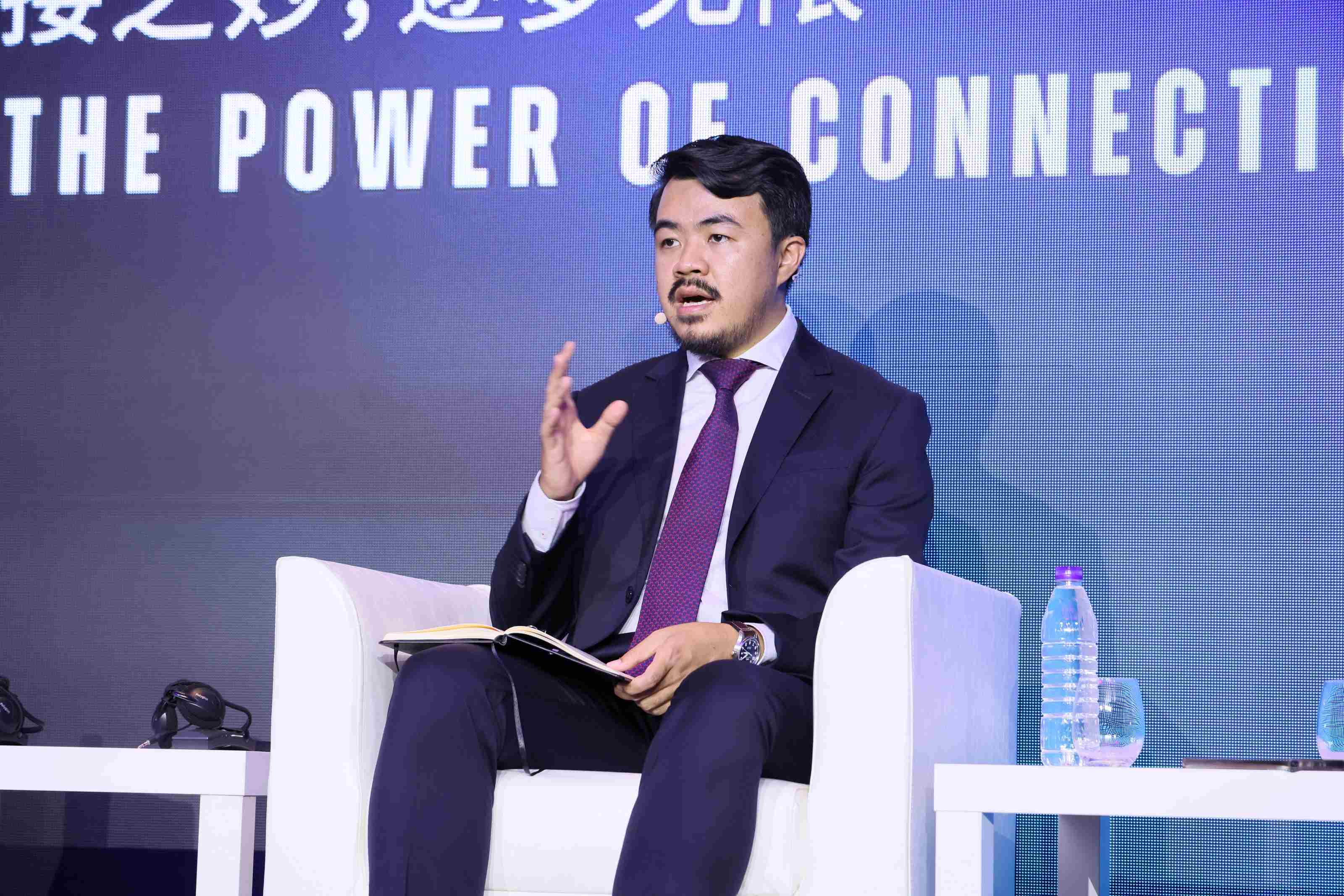 A panelist seated on a white chair, holding a notebook and gesturing mid-discussion.