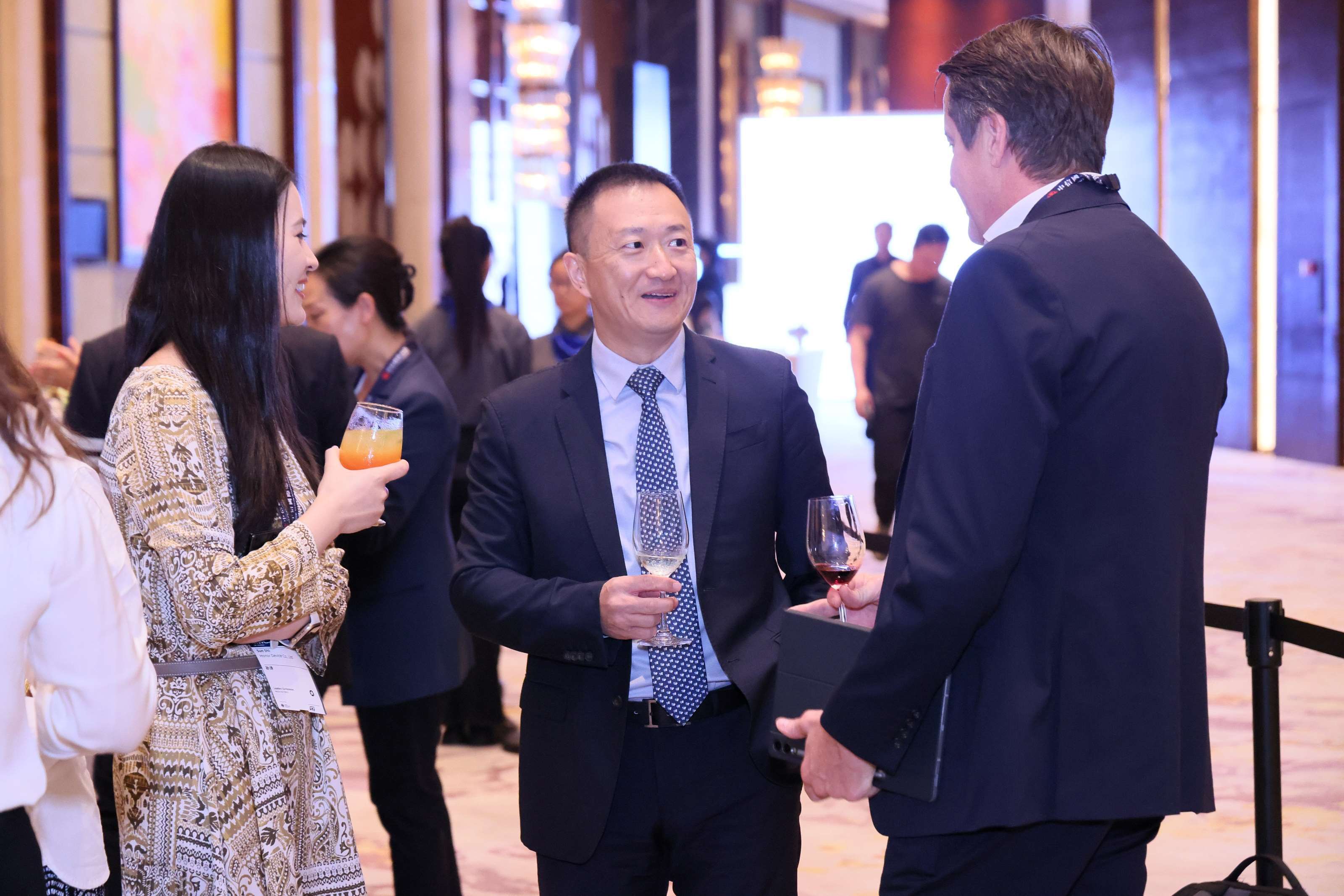 A group of formally dressed individuals conversing at a professional event, with drinks in hand, set against the warm lighting and elegant decor of an indoor venue.
